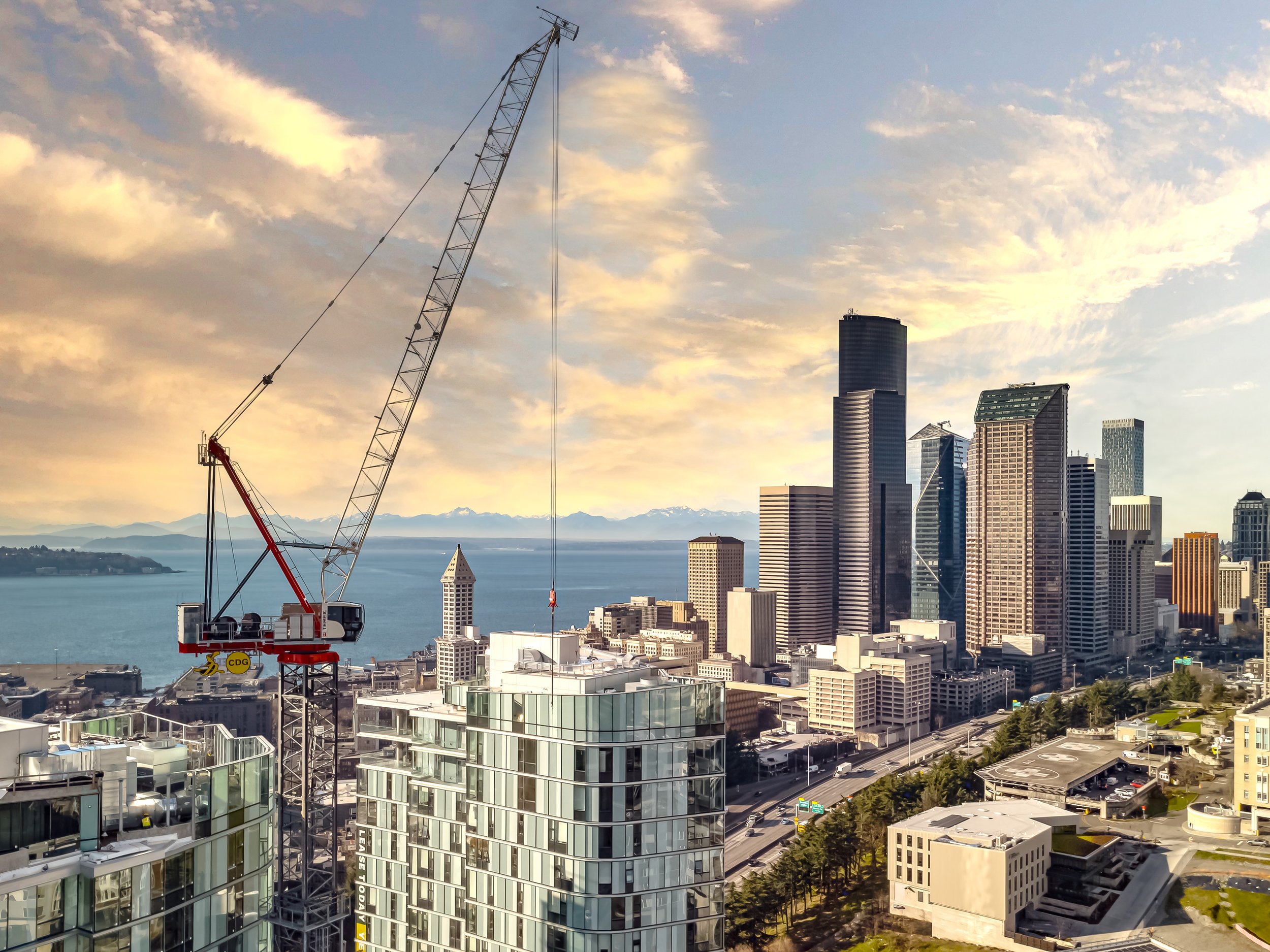 Seattle skyline with tall skyscrapers by the water, a construction crane in the foreground, and mountains in the background under a partly cloudy sky at sunset.