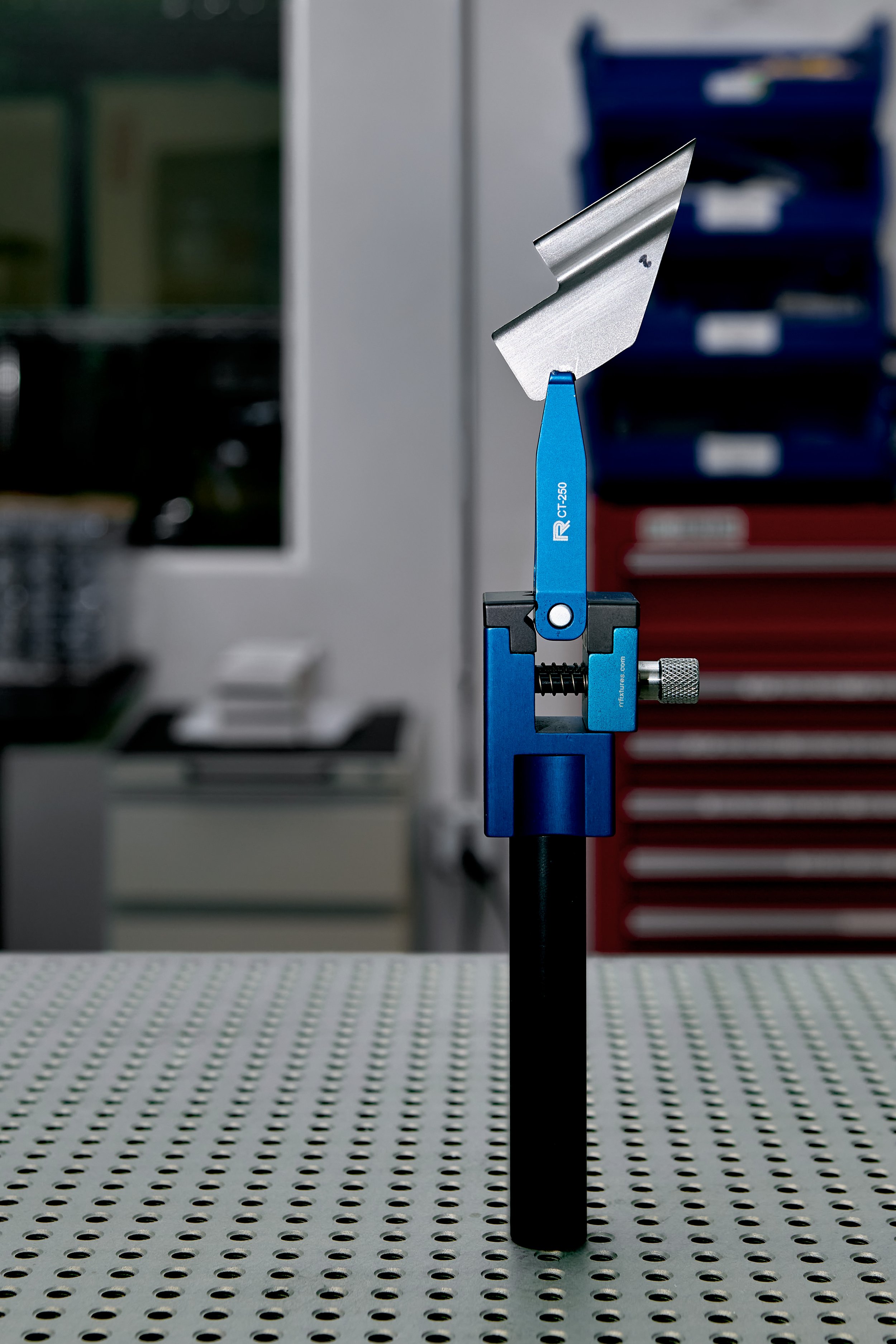 Vertical metal blade attached to a blue measuring fixture, placed on a perforated white table, with a blurred workshop background.