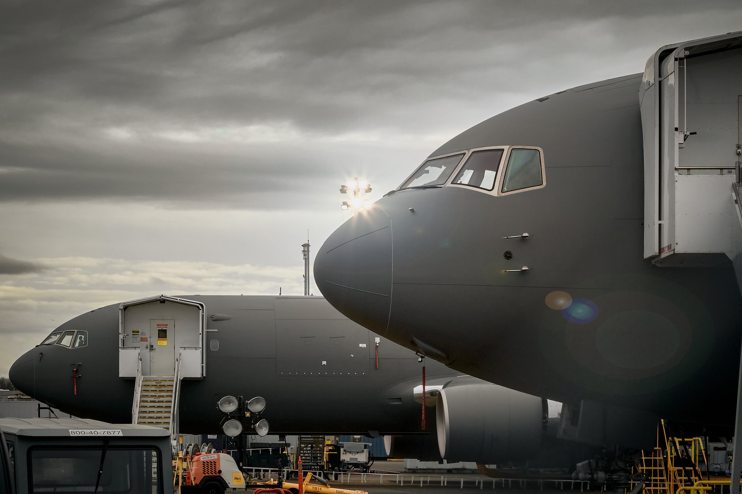 Two large military cargo planes with gray bodies parked on an airport tarmac under a cloudy sky.