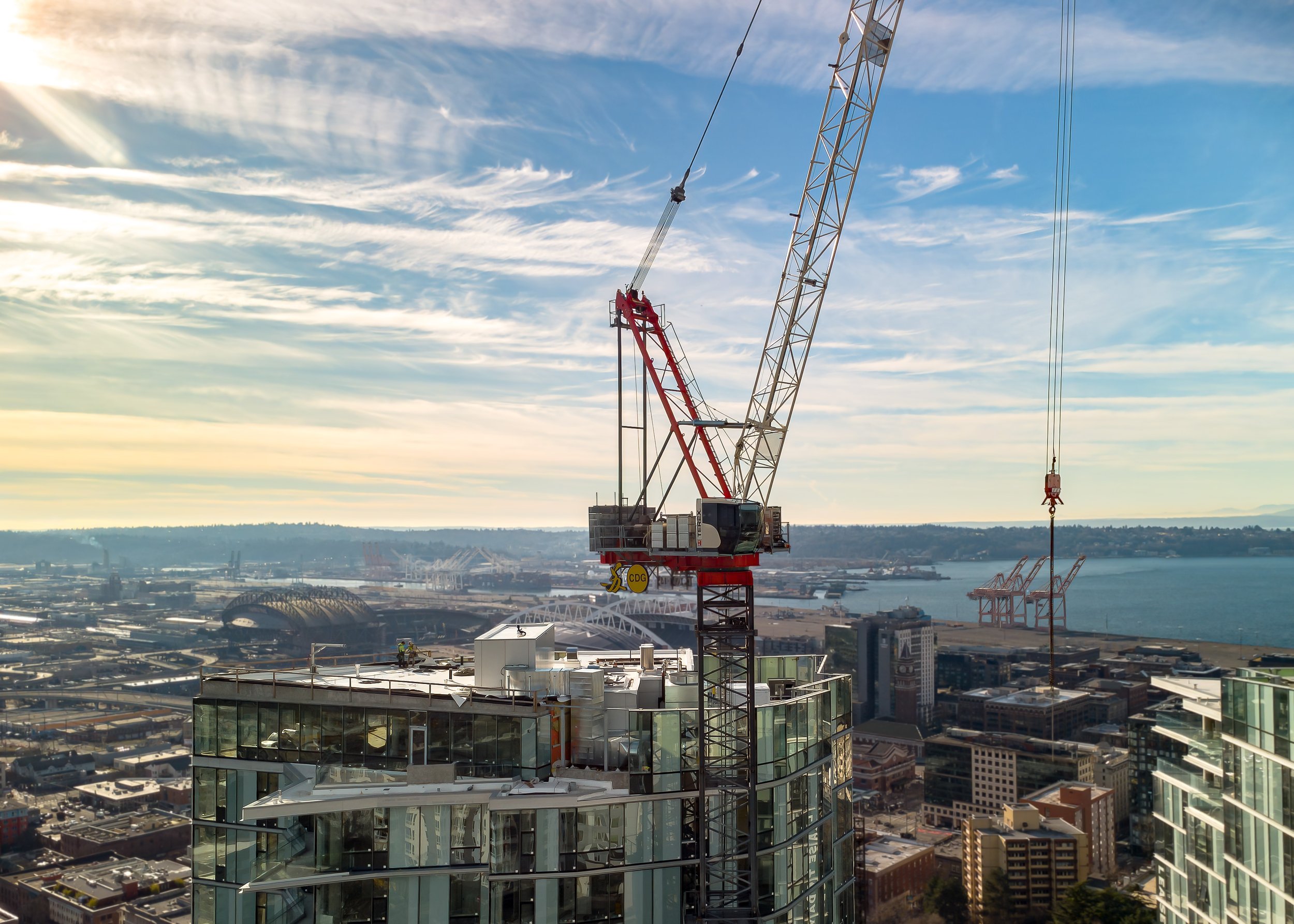 A construction crane positioned on top of a tall building under a partly cloudy sky, overlooking a cityscape and harbor.