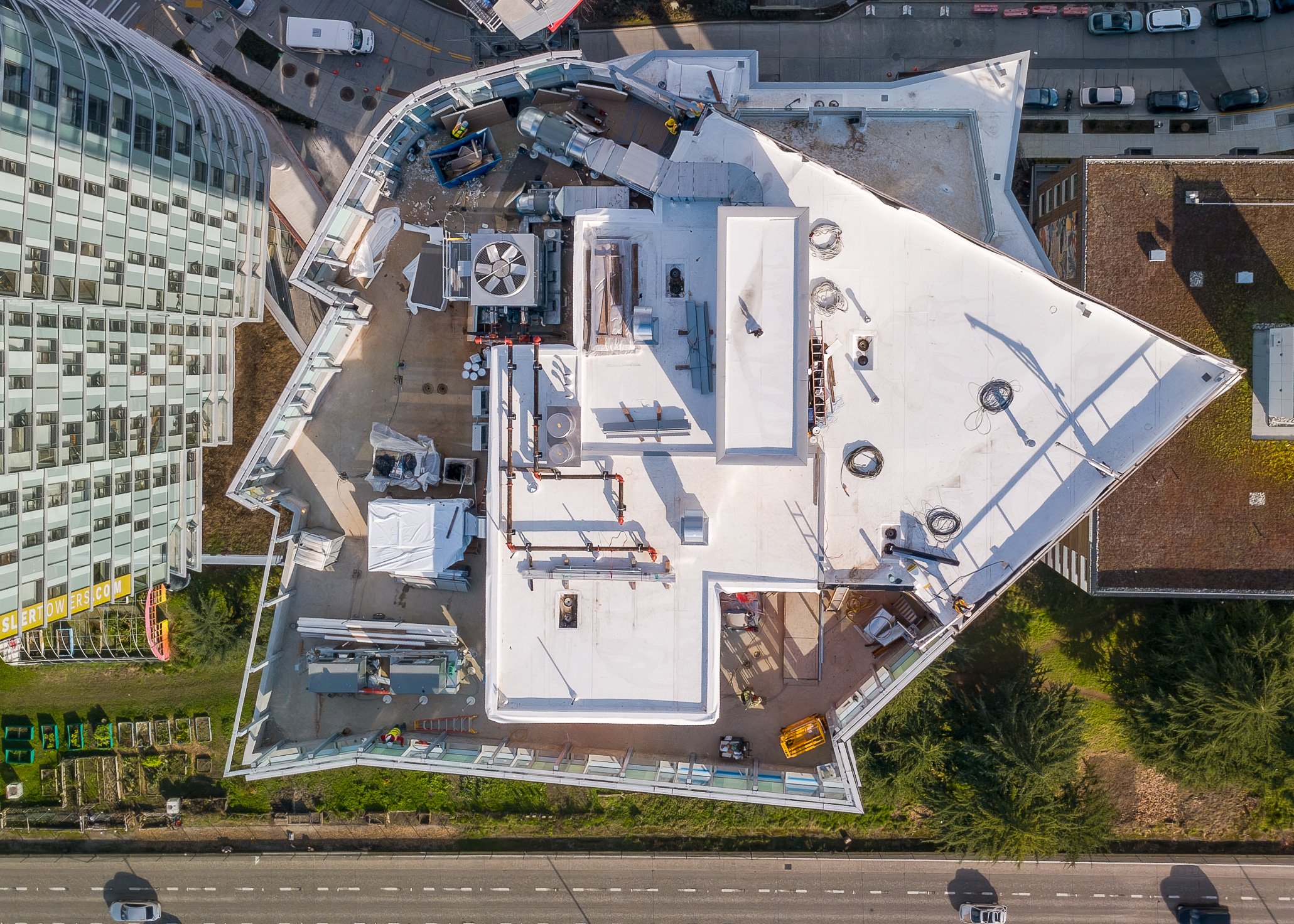 An aerial view of a building rooftop under construction, with various equipment, vents, and a crew members working, surrounded by streets and nearby buildings.