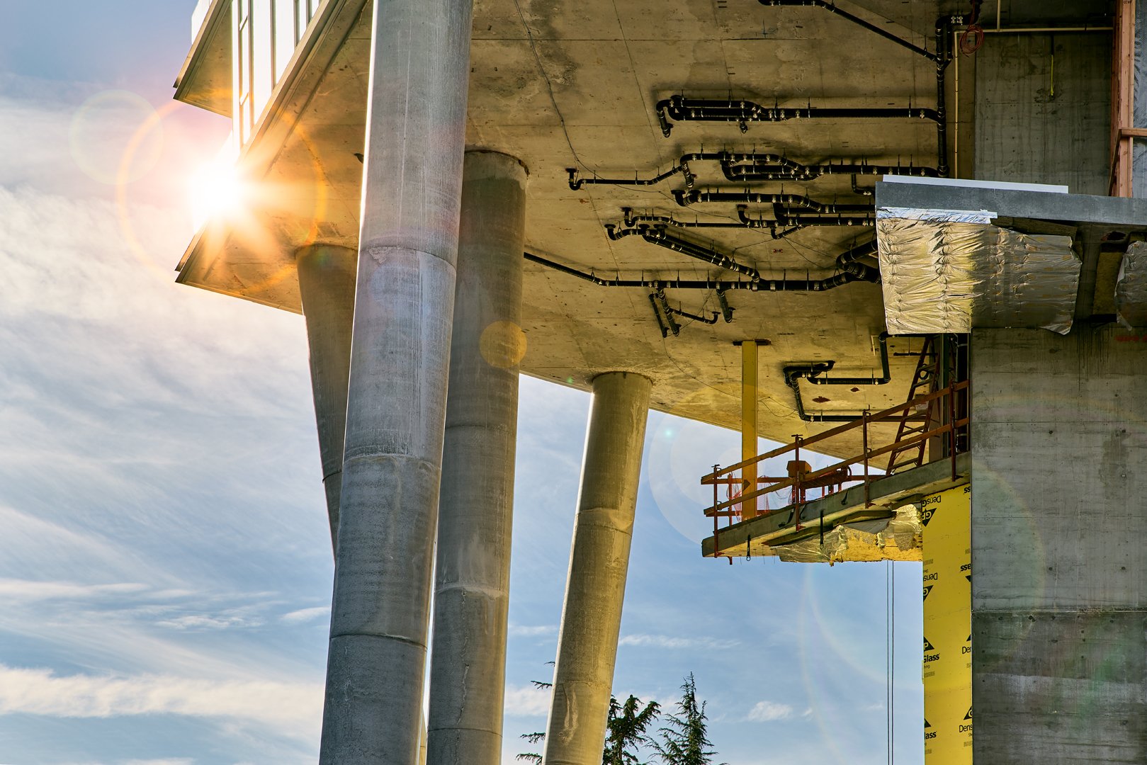 Construction site showing the underside of a building with large support columns, visible pipes, and partially installed insulation panels. The sun is shining, creating lens flares in the image.
