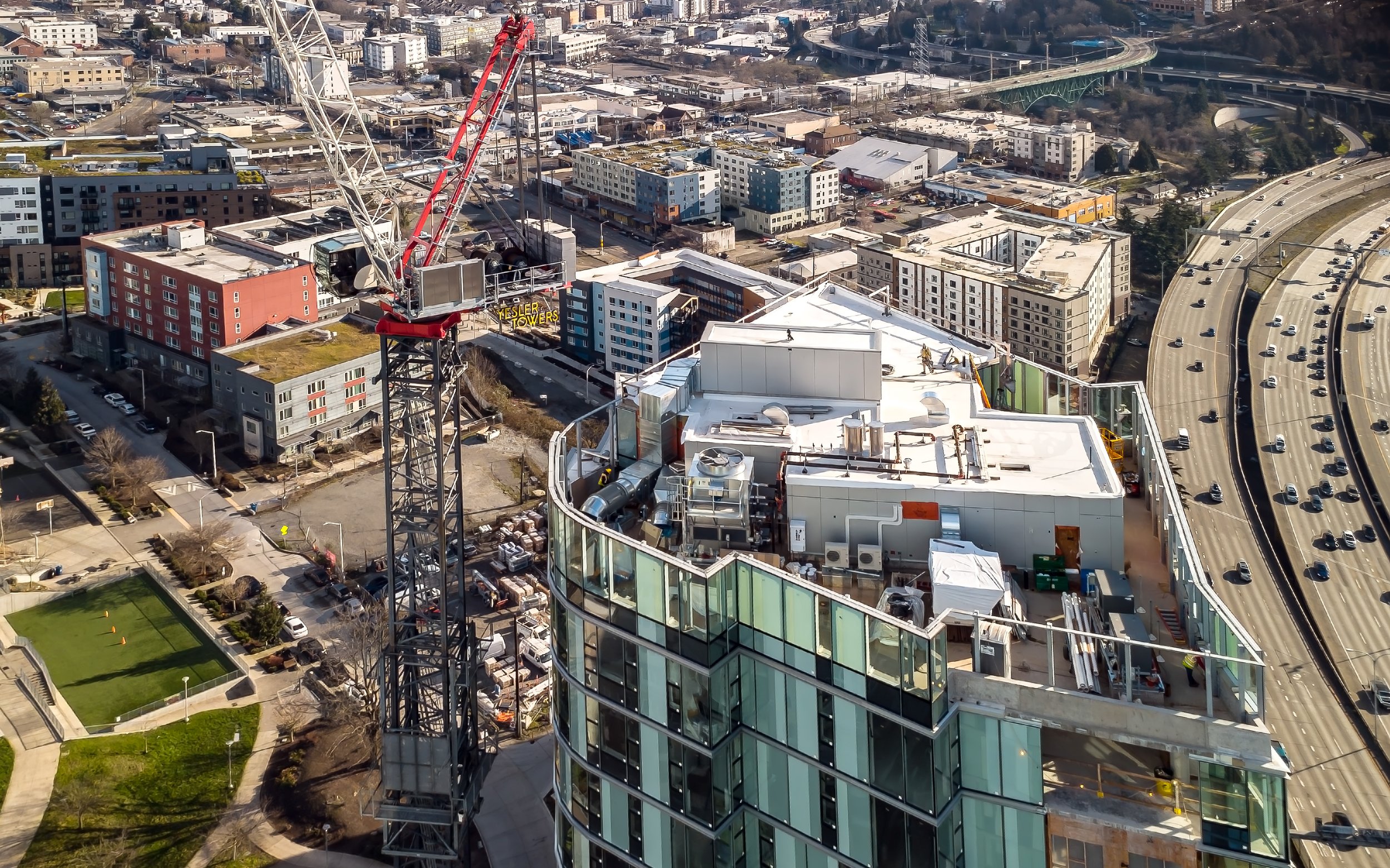 Aerial view of a cityscape showing a high-rise building under construction with a large crane on top, surrounded by roads, parking lots, and other buildings.