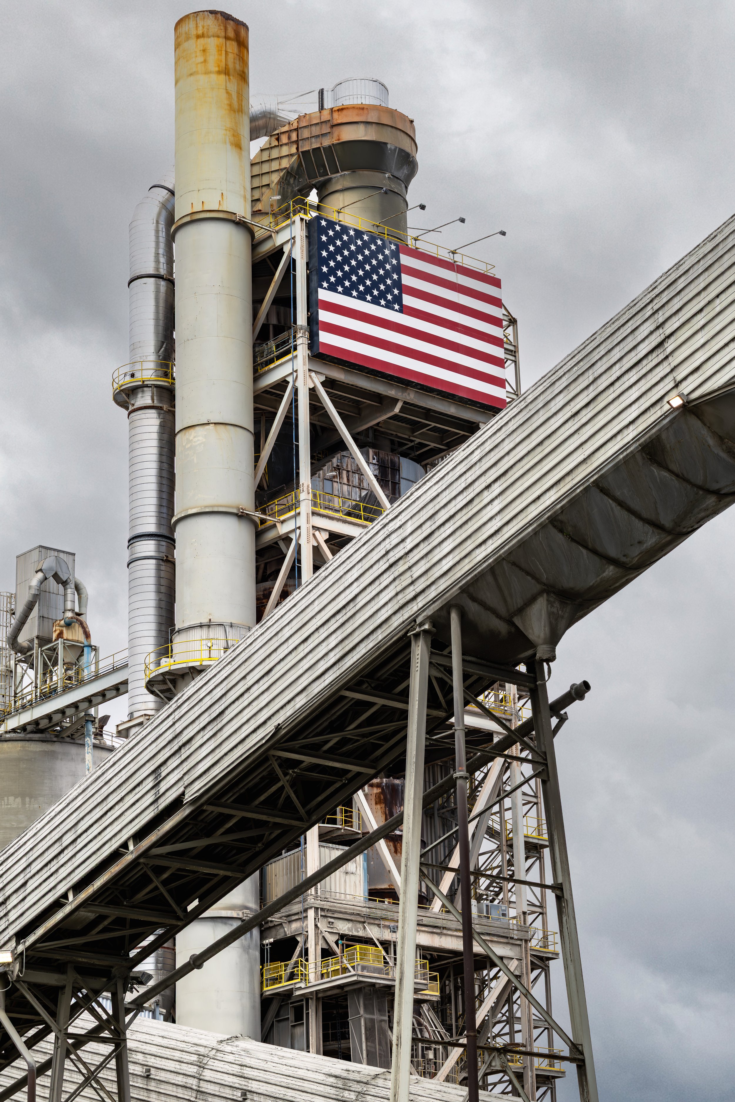 Industrial factory with towering metal structures, pipes, and yellow safety railings, displaying an American flag.