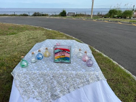 Table with decorative glass jars and a colorful sand art frame, set outdoors with a road and lake view in the background.