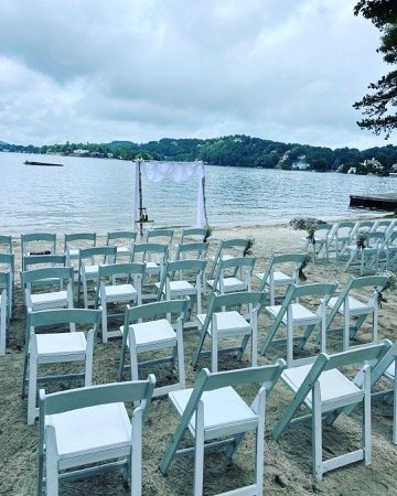 Beach wedding setup with rows of white chairs facing an archway by the water on a cloudy day.