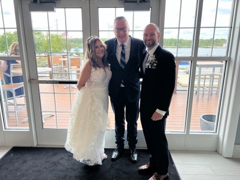 Bride and groom posing with a man in a suit by a glass door with outdoor seating in the background.
