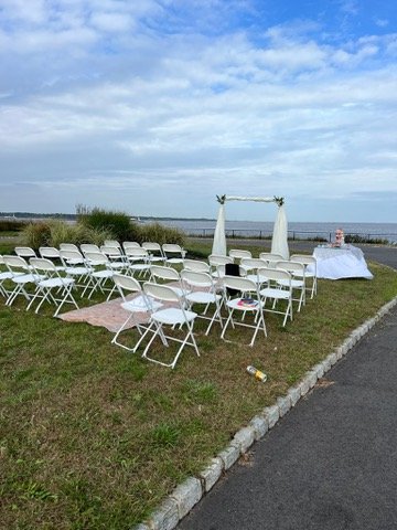 Outdoor wedding setup with white chairs facing a small arch near the ocean, cloudy sky and grassy area.