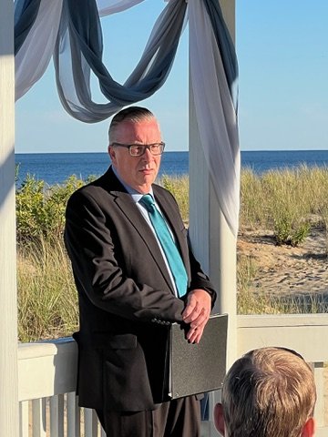 Man in a suit standing at a beachside venue, holding a folder, with ocean and sand dunes in the background. Decor includes draped fabric.