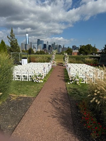 Outdoor wedding ceremony setup with white chairs and floral decorations, overlooking a city skyline under a partly cloudy sky.