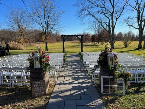 Outdoor wedding setup with white chairs arranged in rows facing a wooden arbor. The pathway is lined with floral decorations on wooden barrels. Trees and a grassy field are visible in the background under a clear blue sky.