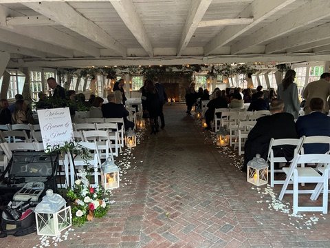 Indoor wedding aisle with white chairs, surrounded by decorative lanterns and floral arrangements, guests seated, brick floor, and floral archway at the end.