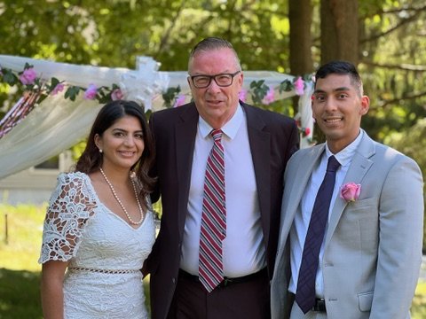 Three people posing outdoors, with a wedding arch decorated with flowers and tulle in the background. The person in the center is wearing a suit and glasses, while the two on the sides are wearing wedding attire; the one on the left is in a lace dres