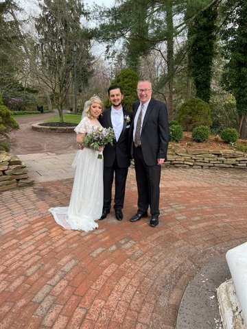 A bride in a white dress and groom in a black suit stand next to an older man in a suit on a brick path, with trees and bushes in the background.