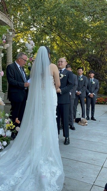 Bride and groom exchanging vows outdoors with officiant and groomsmen, surrounded by floral decorations.