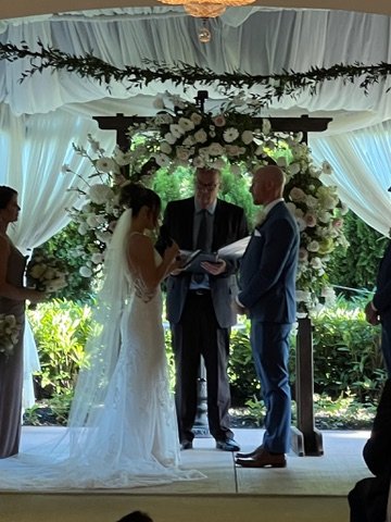 Bride and groom stand facing each other under a floral arch during a wedding ceremony, with an officiant and bridesmaid present.