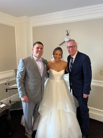 Bride in a strapless white wedding gown standing with two men, one in a light gray suit with a bow tie and the other in a dark suit, in an elegantly decorated room.