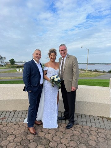Three people in formal attire standing outdoors, with a woman in a white dress holding a bouquet, flanked by two men in suits.