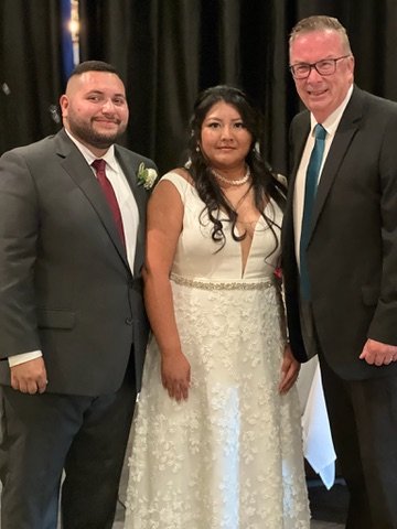 Three people posing together at a formal event, with one wearing a wedding dress and the other two in suits.