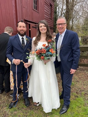 A bride and groom in wedding attire stand with a third man in suits, smiling outdoors in front of a red wooden building. The bride is in a white dress holding a bouquet, and the groom and the third man wear suits.