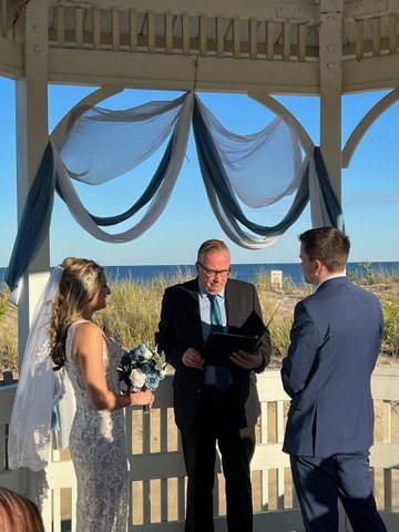 A wedding ceremony by the beach with a bride in a white dress and veil, a groom in a blue suit, and an officiant in a suit standing under a gazebo decorated with white and blue drapery.