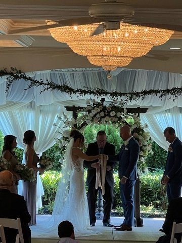 Wedding ceremony under a floral arch with the bride and groom holding hands, surrounded by a bridal party. Elegant chandelier overhead, white drapery and greenery decor.