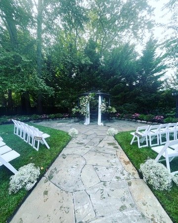 Outdoor wedding setting with a stone path leading to a flower-adorned arch, surrounded by white chairs and greenery.