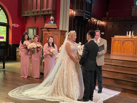 Bride and groom standing at an altar exchanging vows, with bridesmaids in pink dresses holding bouquets, and officiant in the background, in a red and wooden church setting.