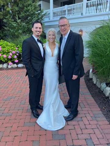 Three people standing outdoors on a brick pathway, with greenery and flowers in the background. One woman in a white gown and two men in suits smile at the camera.