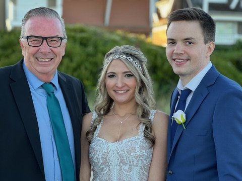 A group photo of three people dressed formally at an outdoor event. A woman in a lace wedding gown stands between two men in suits, one wearing glasses and the other with a boutonniere.
