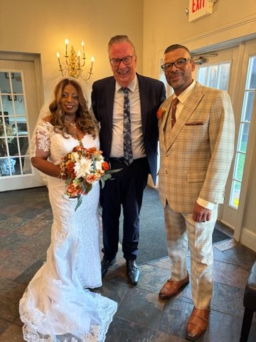 Wedding couple posing with a guest in a formal setting. The bride is wearing a white lace gown and holding a bouquet, while the groom is in a plaid suit. The guest is dressed in a dark suit.