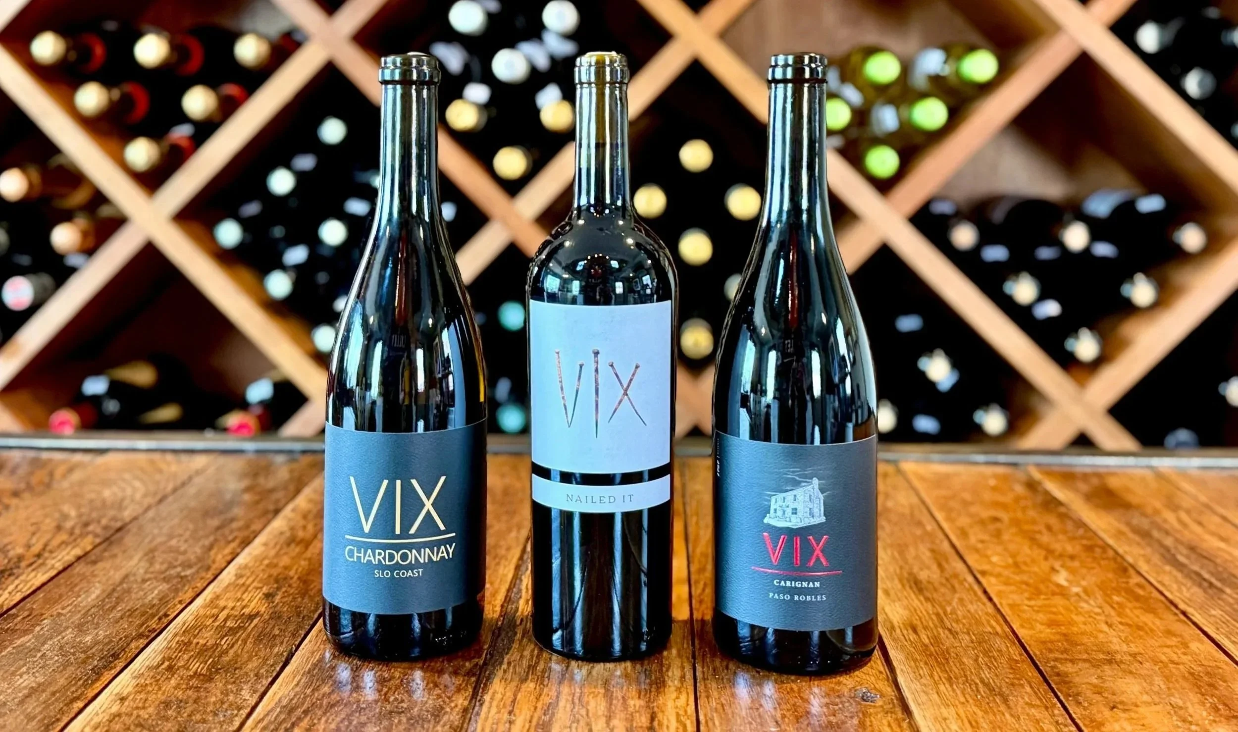 Three bottles of VIX wine placed on a wooden table, with a wine rack full of bottles in the background.