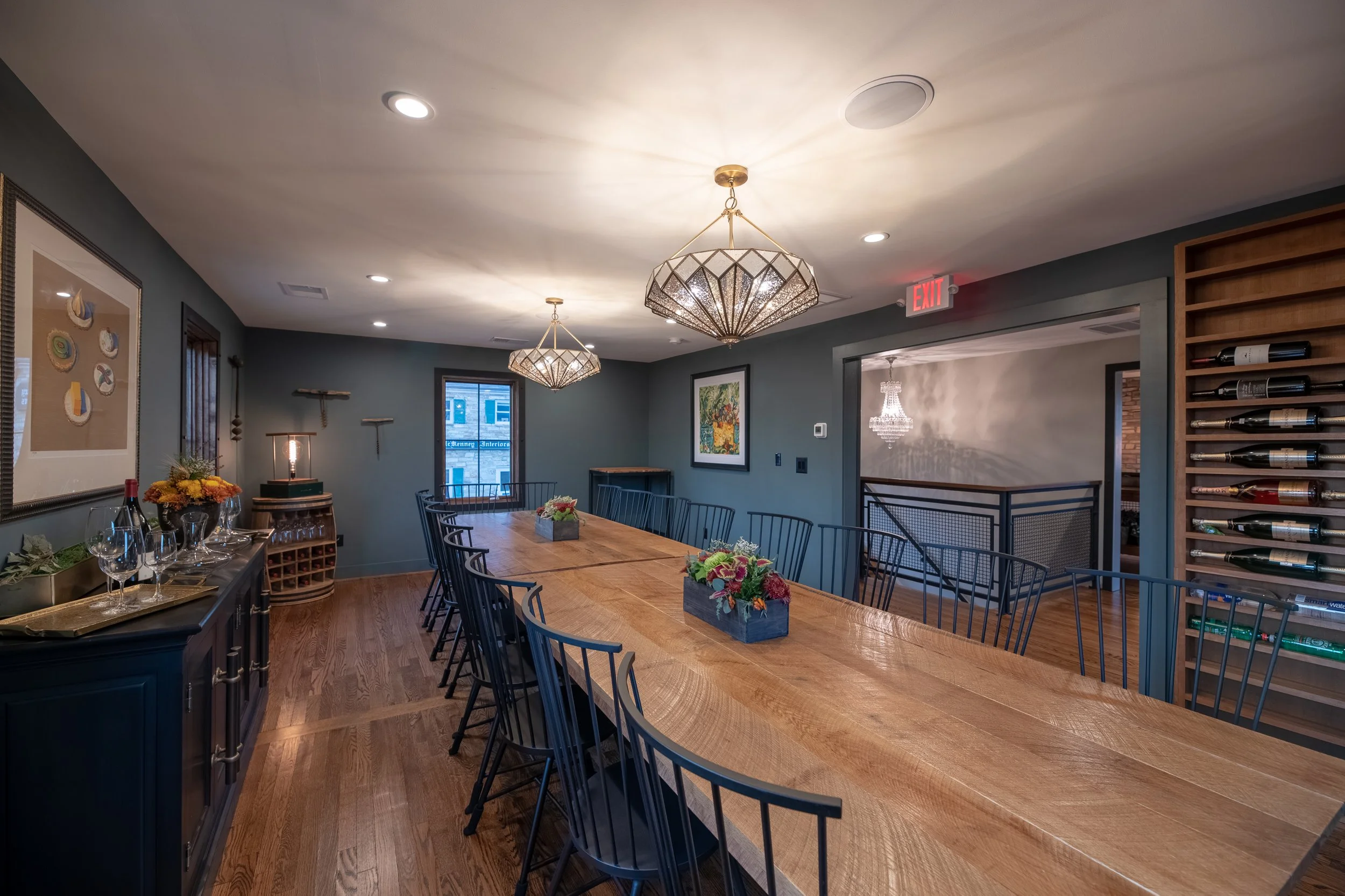 A dining room with a long wooden table with floral centerpieces, black chairs, a blue sideboard with wine glasses and a wine bottle, wall art, two cluster chandeliers, a window, and a staircase with a chandelier.