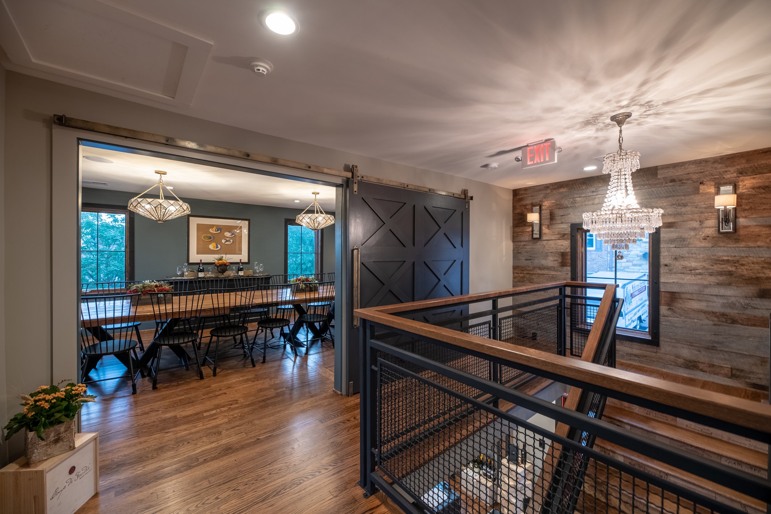 Interior of a modern restaurant or cafe with a wooden staircase, wood-paneled accent wall, chandelier, and a separate dining area with large windows, decorative lighting, and a long table with black chairs.