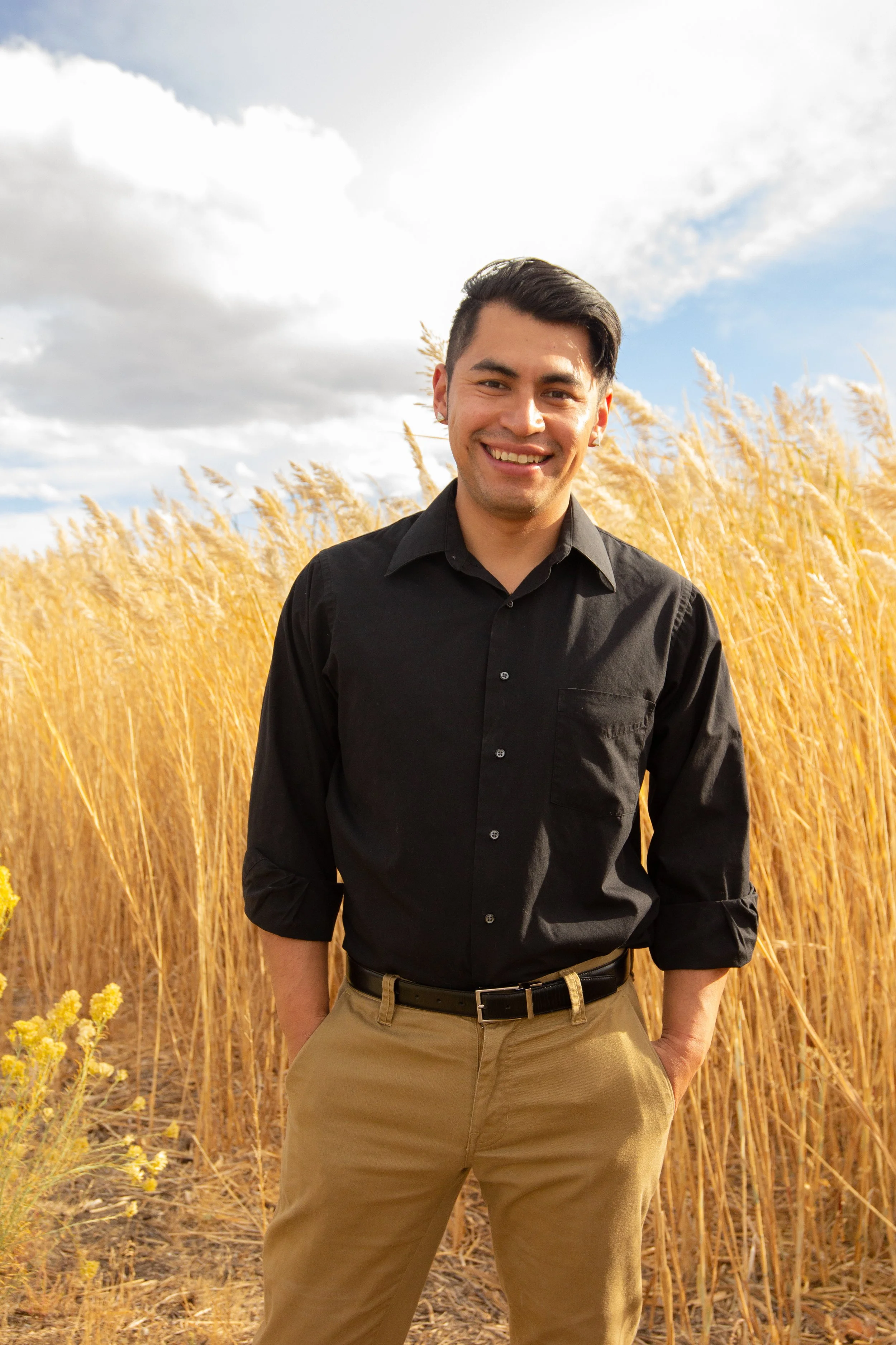 A young man in a black shirt and beige pants smiling outdoors in a field of tall, golden grasses under a partly cloudy sky.