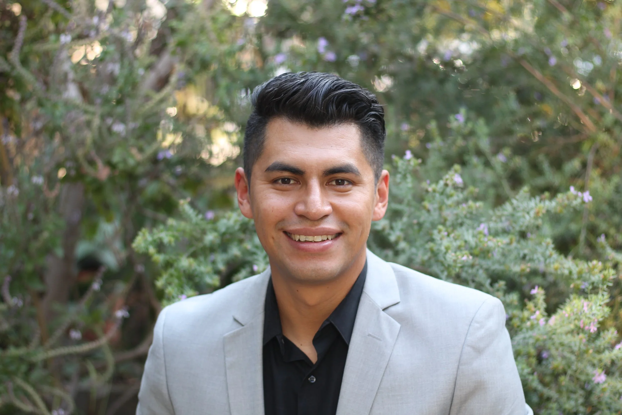 A young man with dark hair and a bright smile, wearing a light gray blazer and black shirt, standing outdoors in front of green bushes and purple flowers.
