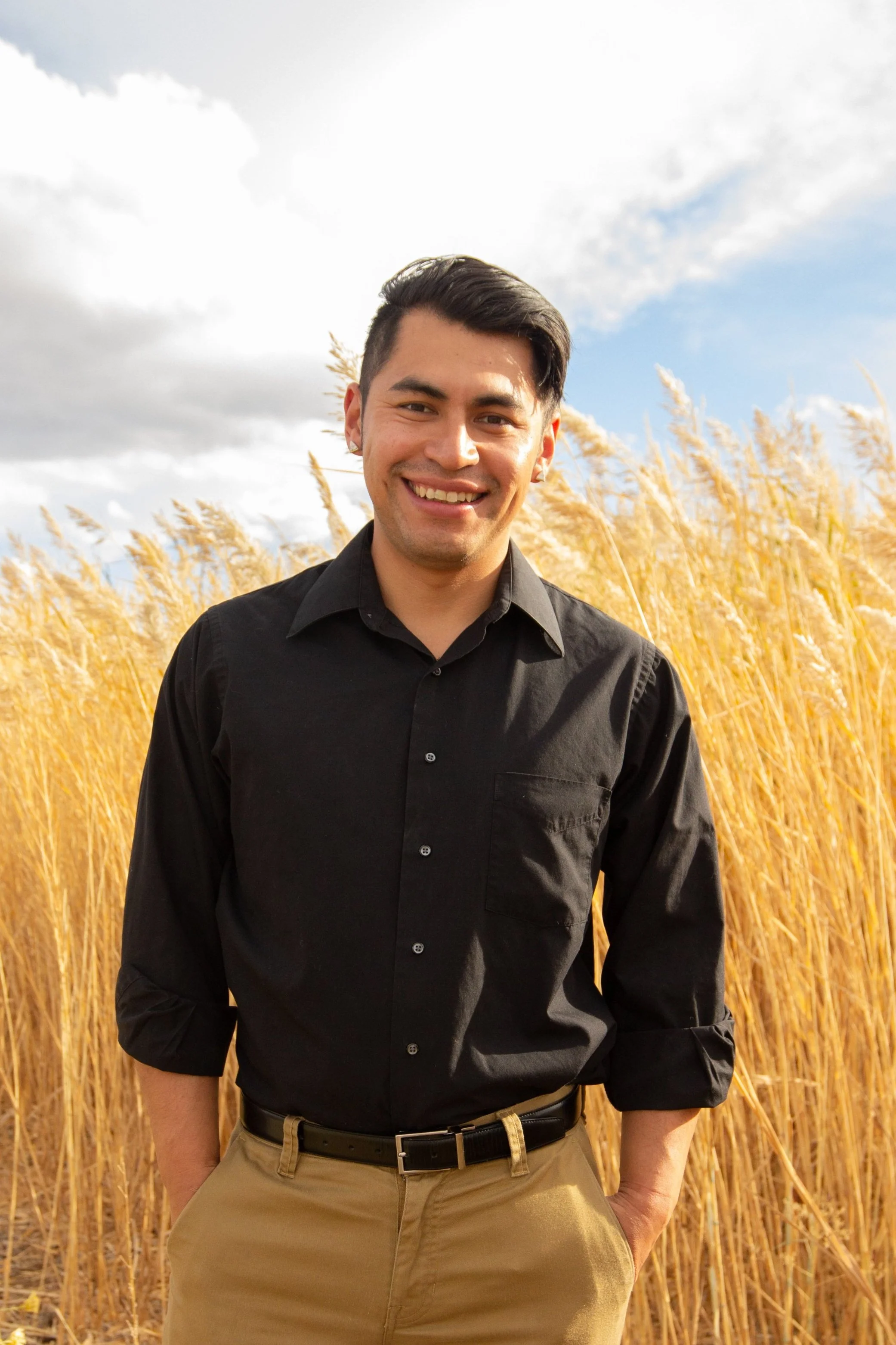 A smiling young man in a black shirt and khaki pants standing in a field of tall, golden grasses on a partly cloudy day.