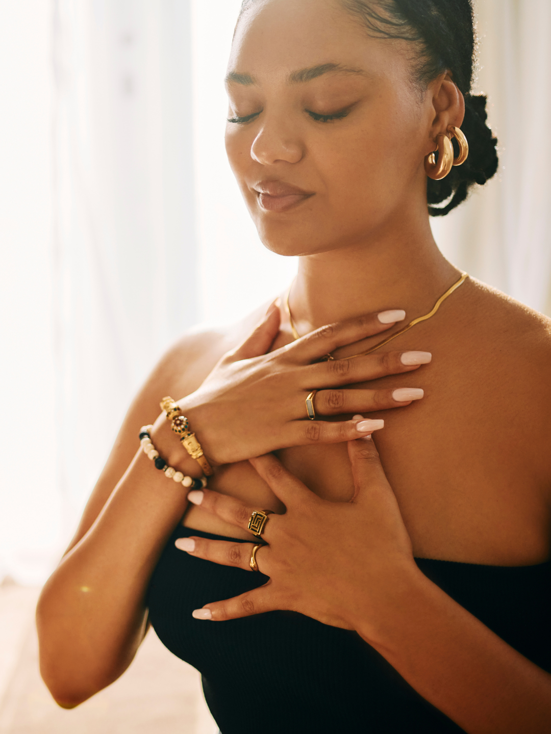 A woman with closed eyes and a gentle smile, wearing gold jewelry including earrings, a necklace, rings, and bracelets, touching her chest with both hands, in a softly lit room.