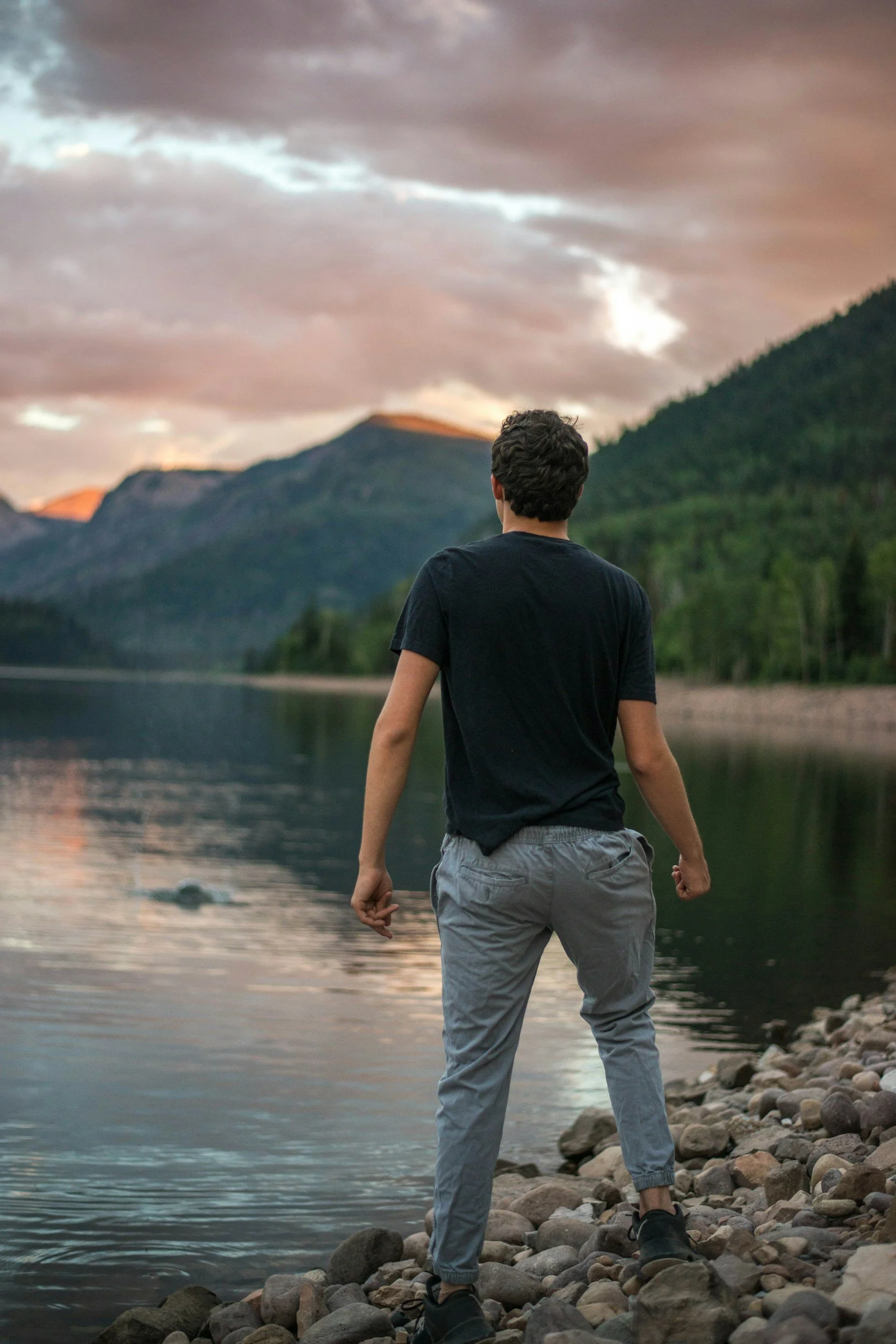 A person stands on a rocky riverbank at sunset, facing a river with mountains and a cloudy sky in the background.