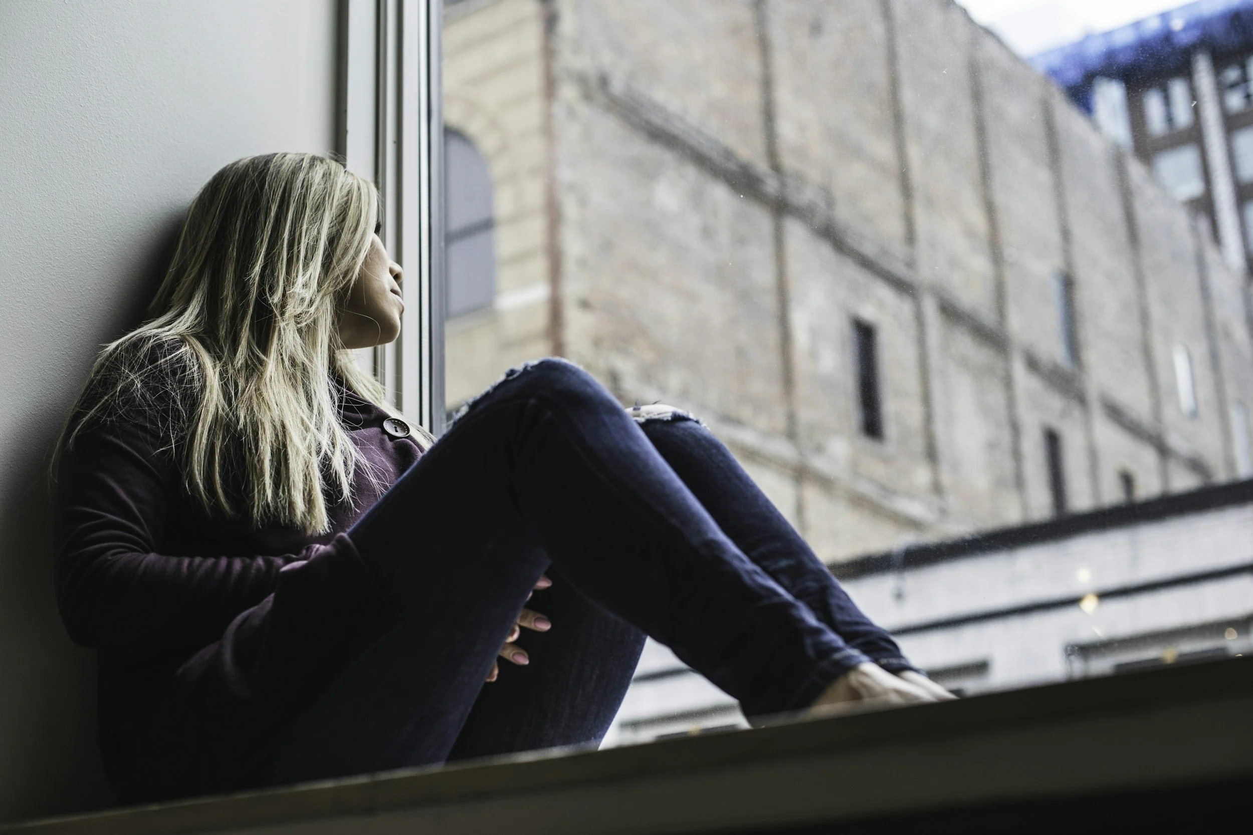 A young woman with blonde hair, wearing a maroon jacket and ripped jeans, sitting by a large window, looking outside at a brick building, relaxed and contemplative.