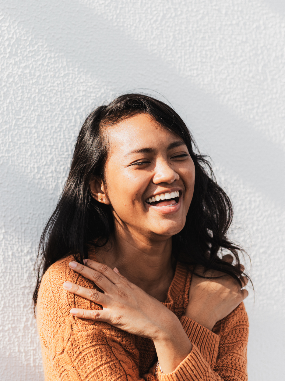 A woman with long dark hair smiling and laughing, wearing an orange sweater, with her hands crossed on her shoulders, standing against a white wall.