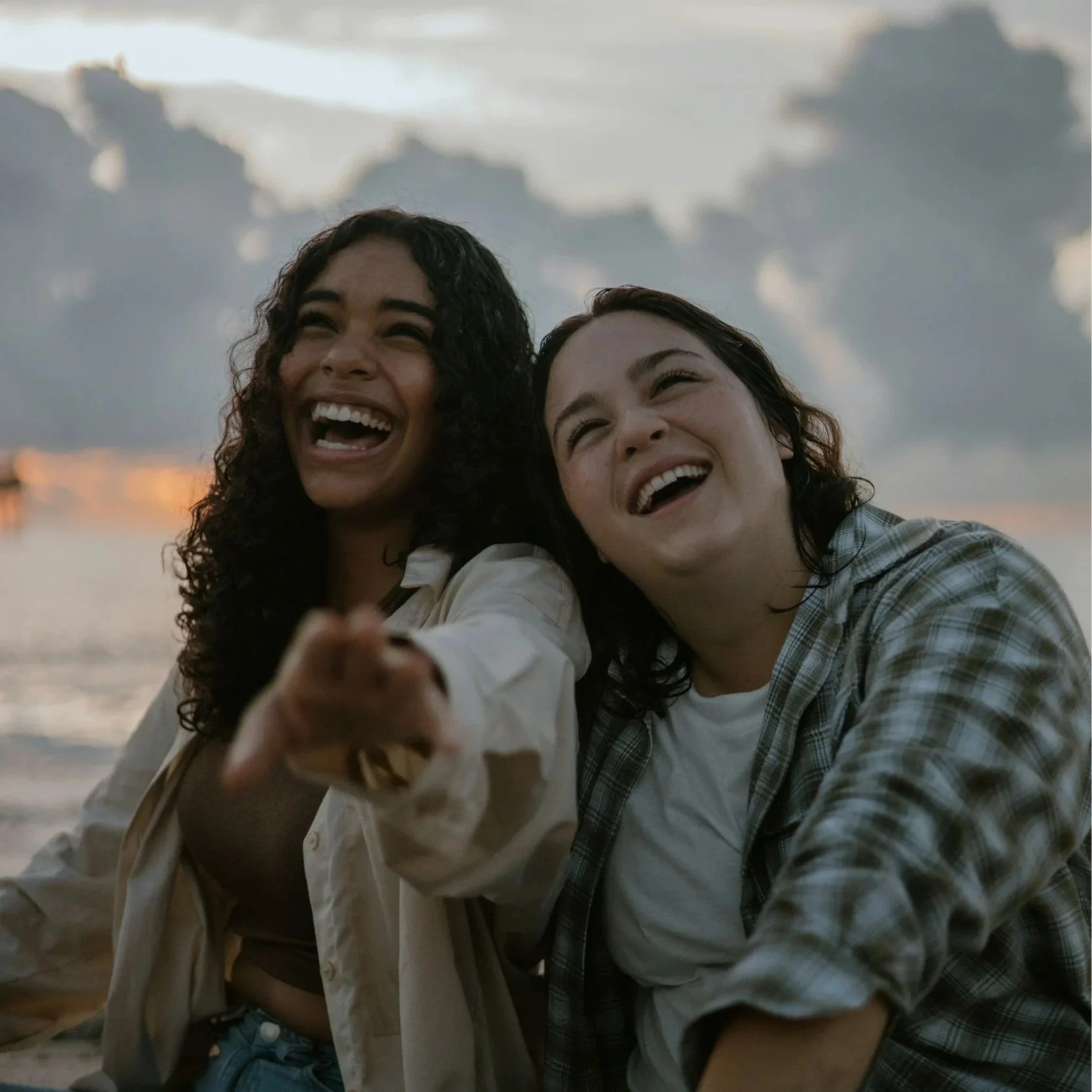 Two women laughing and smiling at sunset on the beach, with ocean and cloudy sky in the background.