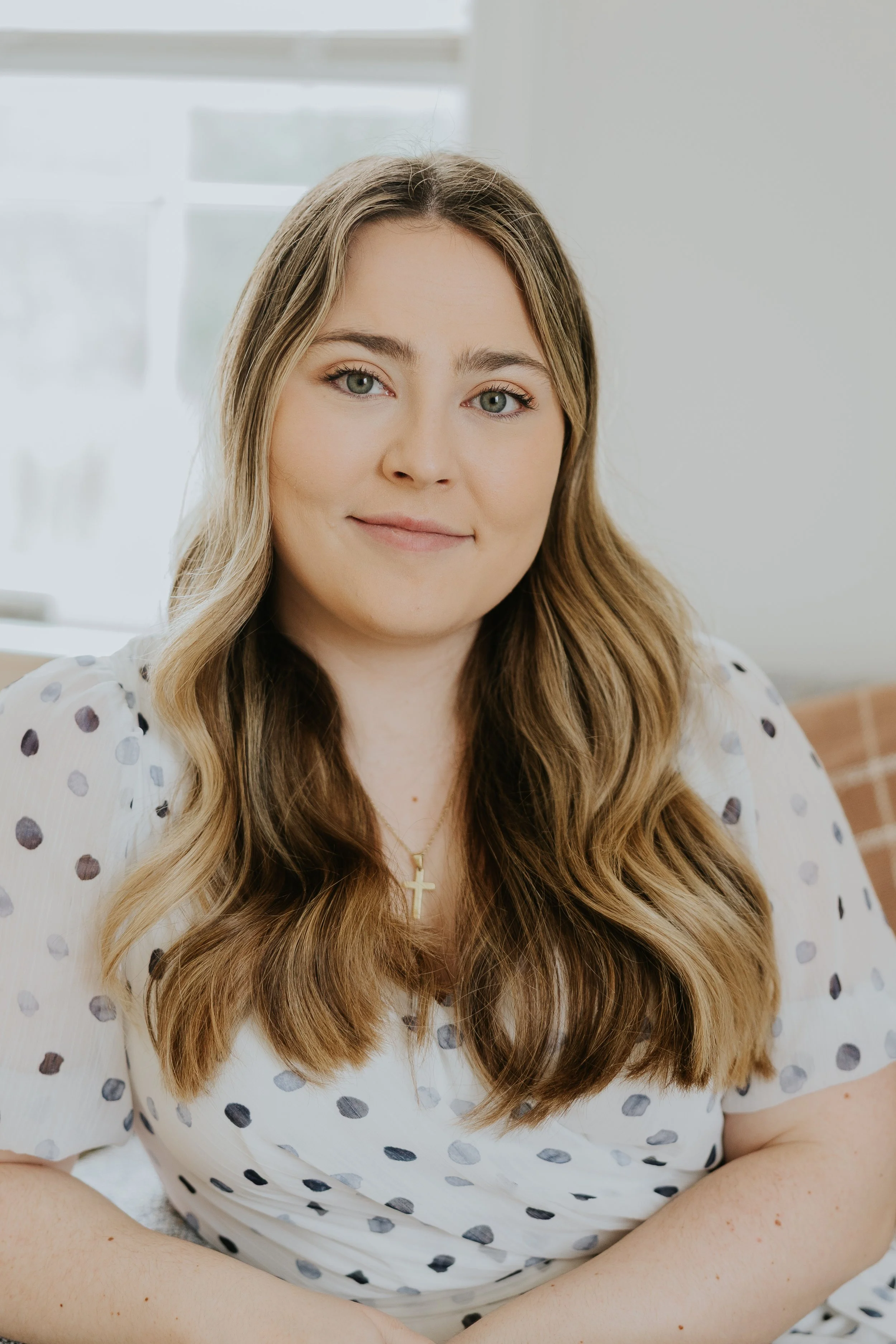 Professional headshot of a young woman with long, wavy brown hair and green eyes, wearing a teal top and a silver necklace with a cross pendant, against a blue gradient background.