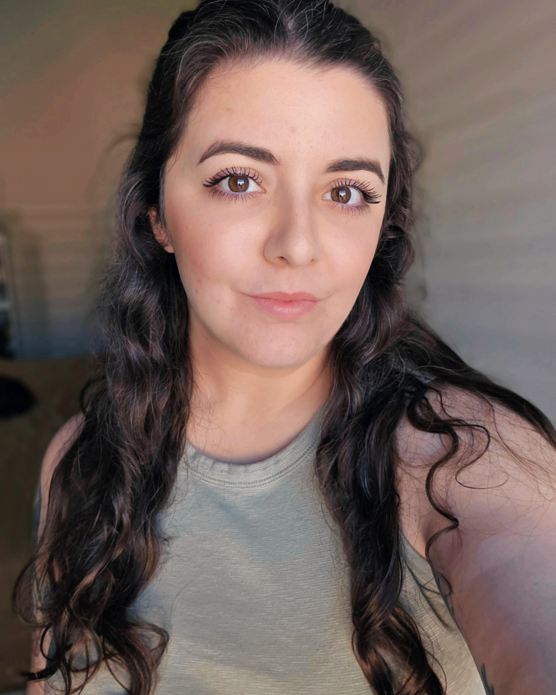 Close-up selfie of author Susan Samadi with long wavy dark hair and brown eyes, wearing a sleeveless light-colored top, indoors with a neutral background.
