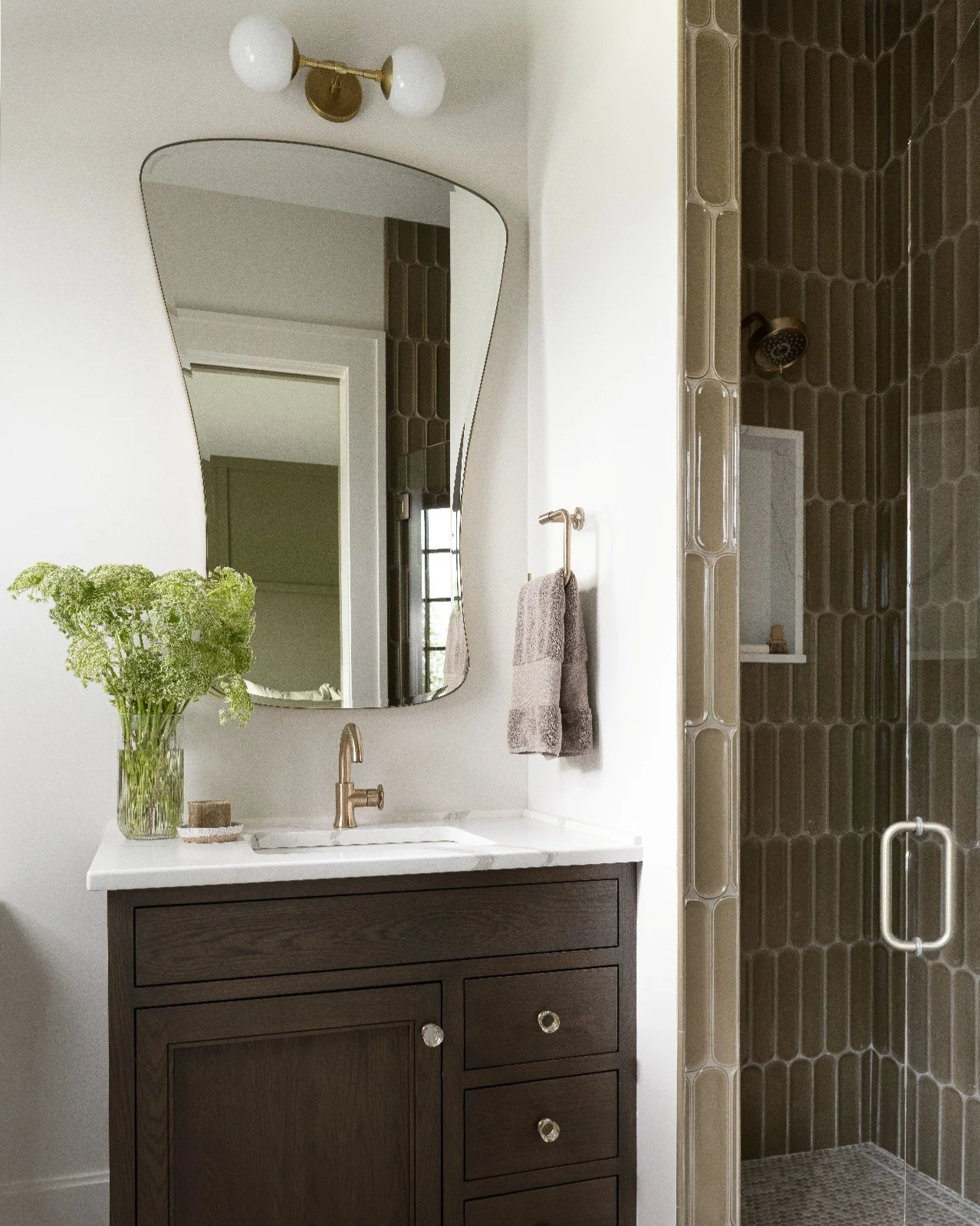 This guest bathroom features rich brown tones, soft brass fixtures, and sculptural tile that subtly echoes the curve of the mirror. It provides a little moment of elevated calm with every visit that is made. 

Builder: @martin.brothers 
Cabinetry: @a