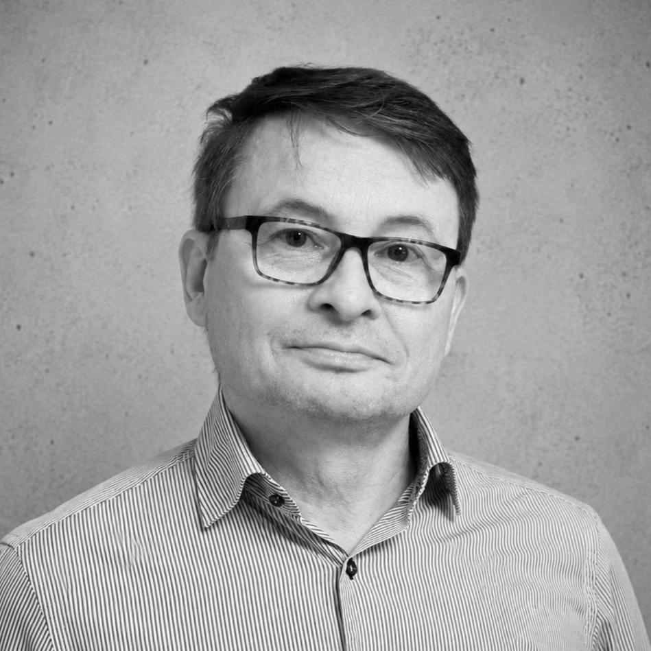 A grayscale image of a person wearing glasses and a striped shirt, leaning against a wall, looking directly at the camera.