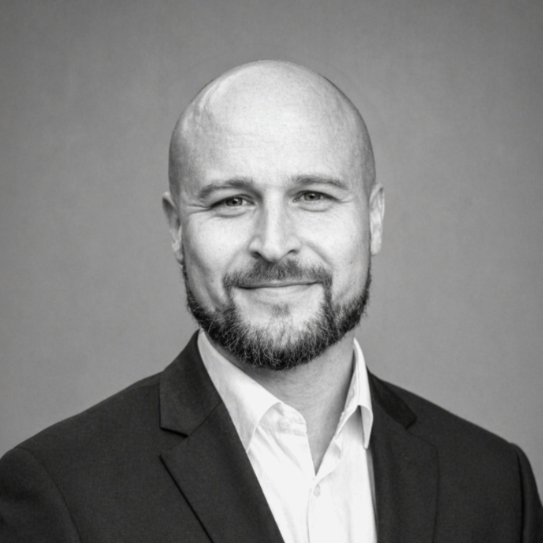 Portrait of a smiling bald man with a beard, wearing a suit jacket and dress shirt, against a gray background.