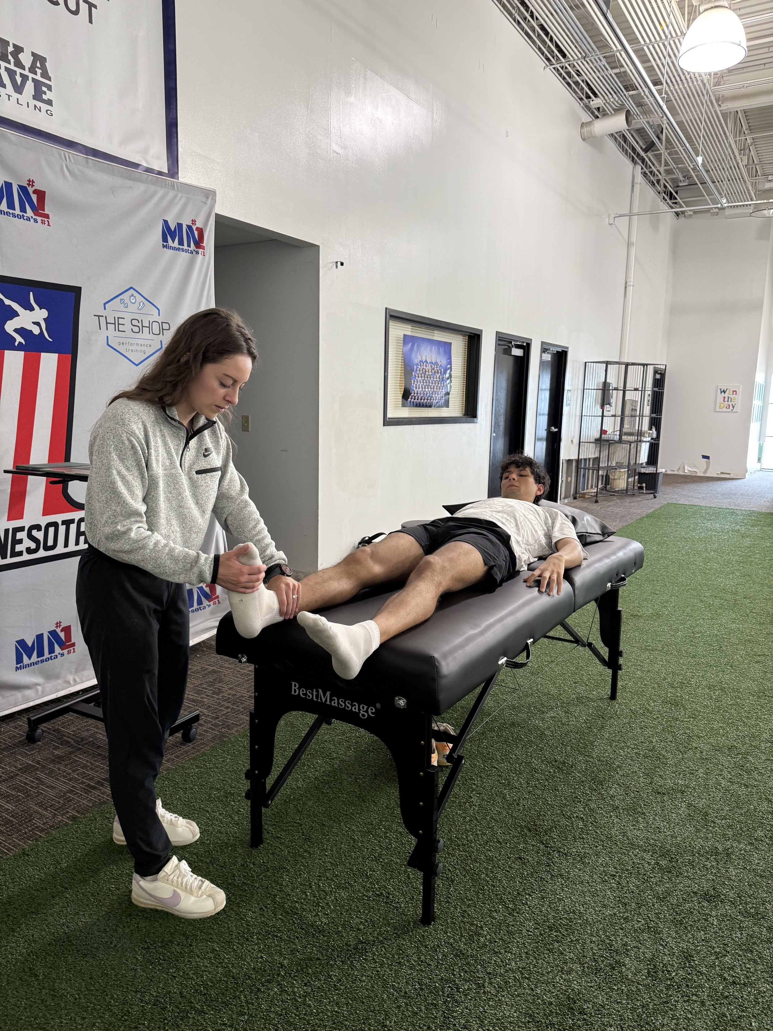 A woman applying a massage or physiotherapy treatment to a man lying on a massage table in a sports facility or therapy center. There are banners and signs on the wall, and a black metal cage on the side.