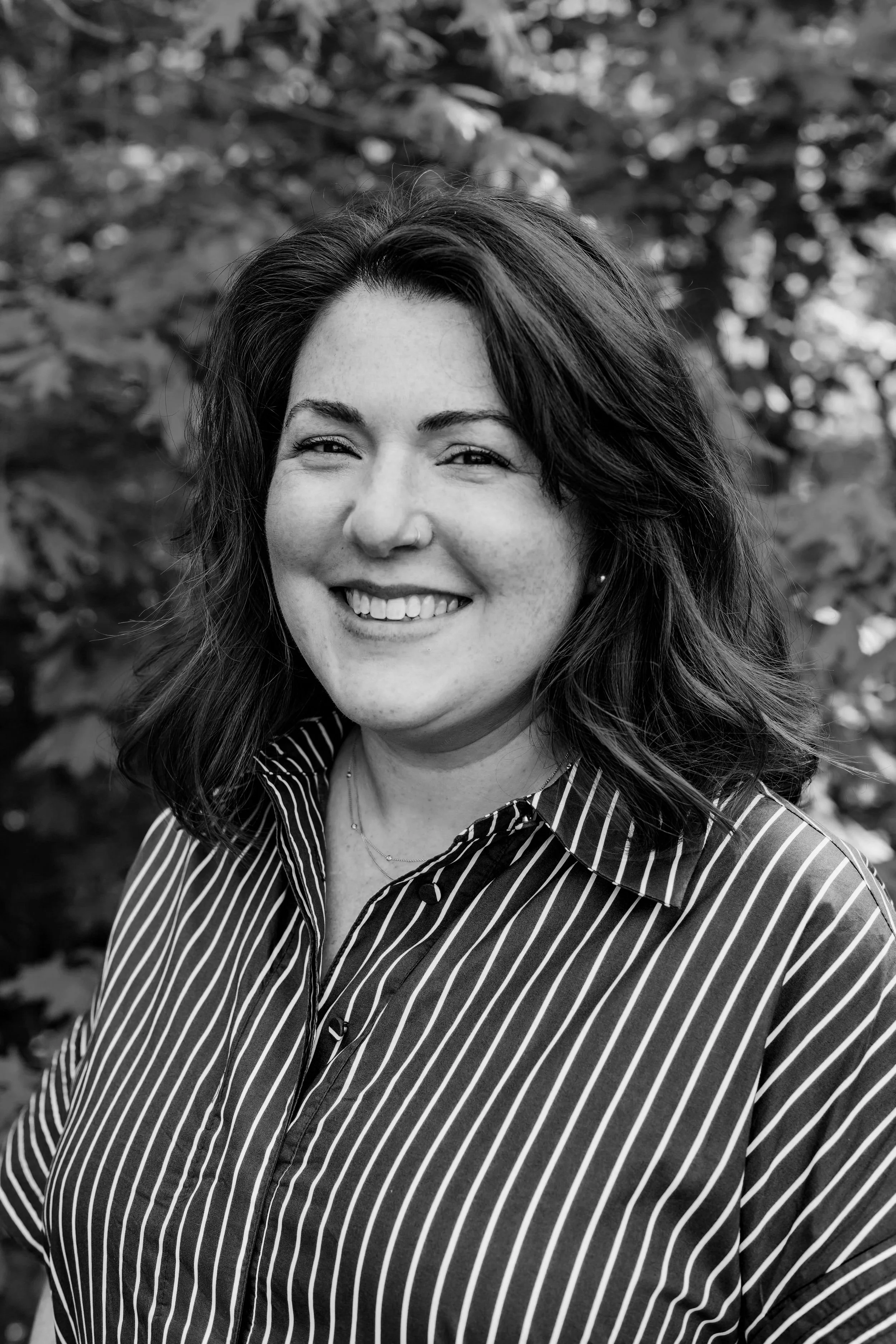A woman with shoulder-length brown hair smiling outdoors in front of green foliage, wearing a green and white striped shirt.