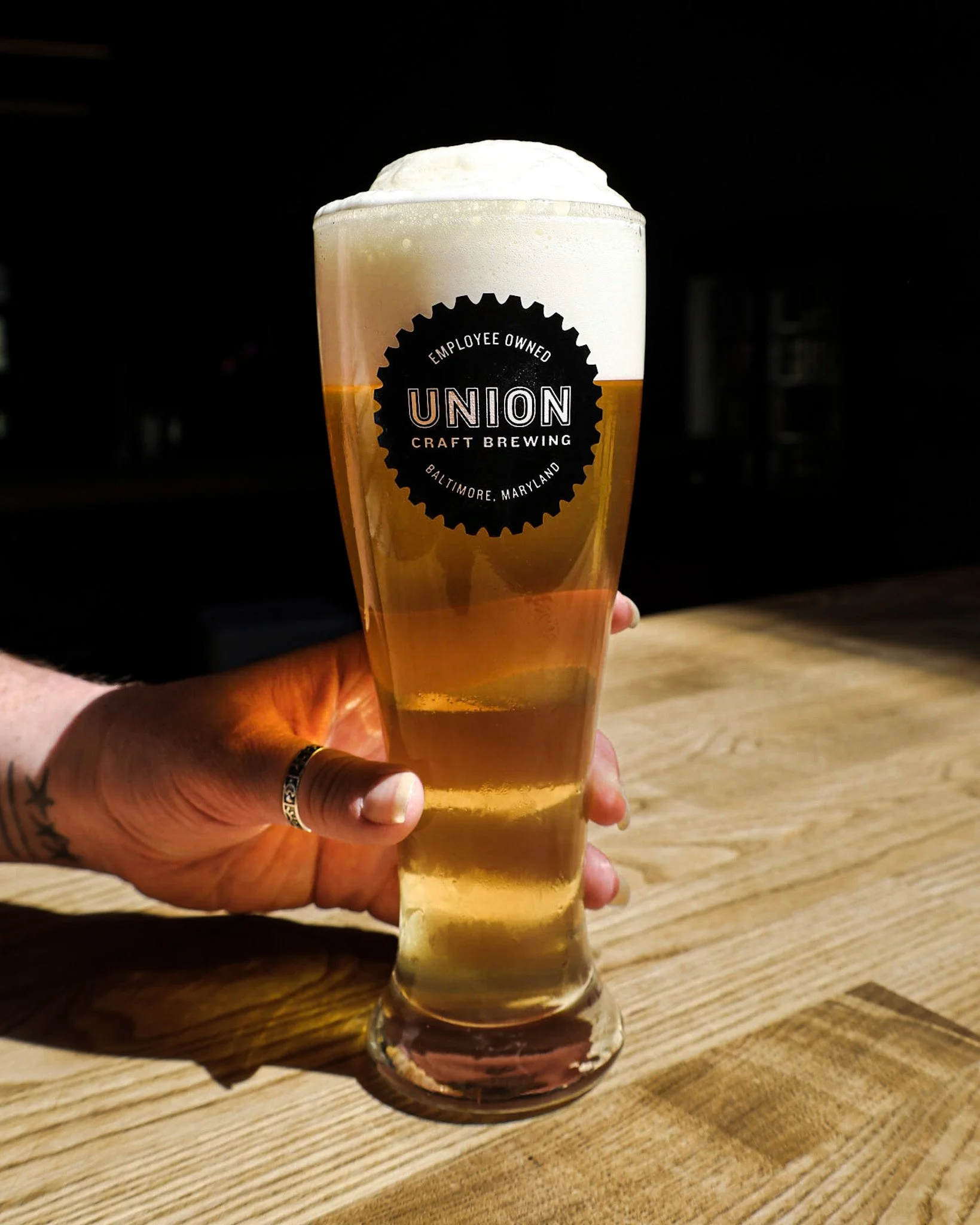 Hand holding a tall glass of beer with a foamy head, logo for Union Craft Brewing in Baltimore, Maryland on the glass, on a wooden table.